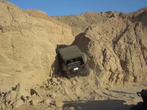 1985 Ford Bronco - A black Jeep Wrangler navigates a challenging off-road course, kicking up dust on a steep sandy descent.