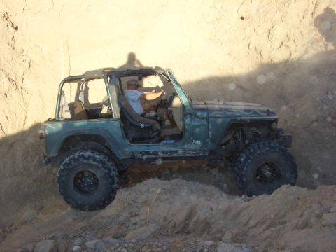 1985 Ford Bronco - A driver navigates a Jeep Wrangler TJ over rough terrain, kicking up dust.