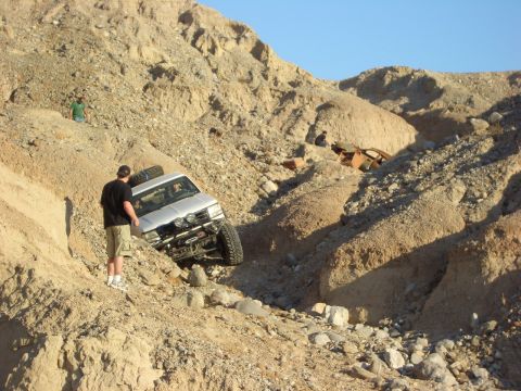1985 Ford Bronco - A modified silver SUV attempts to climb a treacherous, rocky terrain with another vehicle wreck in the distance.