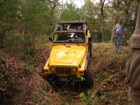 2001 Jeep Wrangler - A yellow 2001 Jeep Wrangler navigates a muddy ditch on a wooded trail, with another vehicle and people in the background.