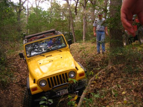 2001 Jeep Wrangler - A yellow 2001 Jeep Wrangler is shown off-roading on a dirt path surrounded by trees.