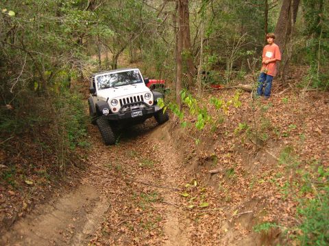 2001 Jeep Wrangler - White Jeep Wrangler driving off-road on a dirt trail with a red vehicle in the background.