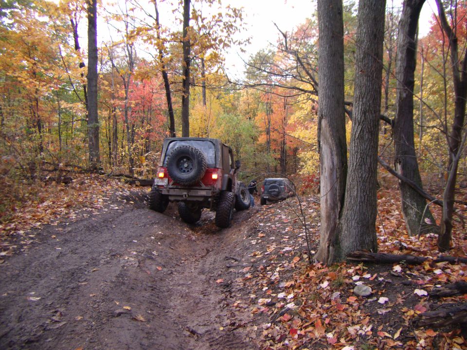 Dark red Jeep Wrangler on a muddy trail, rear view, with another Jeep behind it in autumn woods