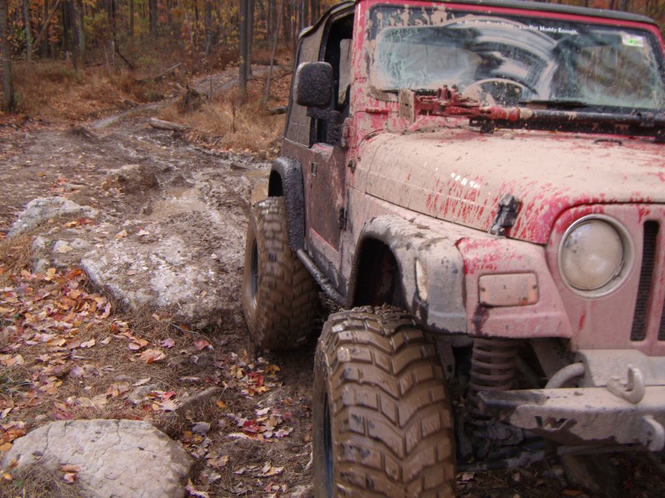 Mud-covered red Jeep Wrangler on a rocky, wooded trail