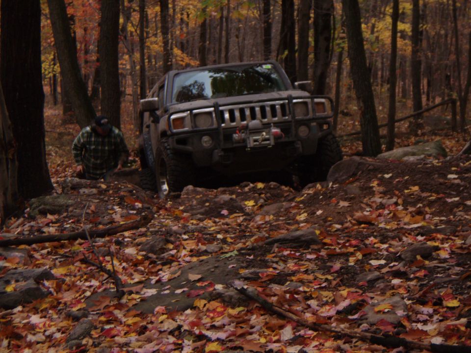 Hummer H3 navigating a rocky, wooded trail covered in autumn leaves. A person in a plaid shirt is visible near the vehicle
