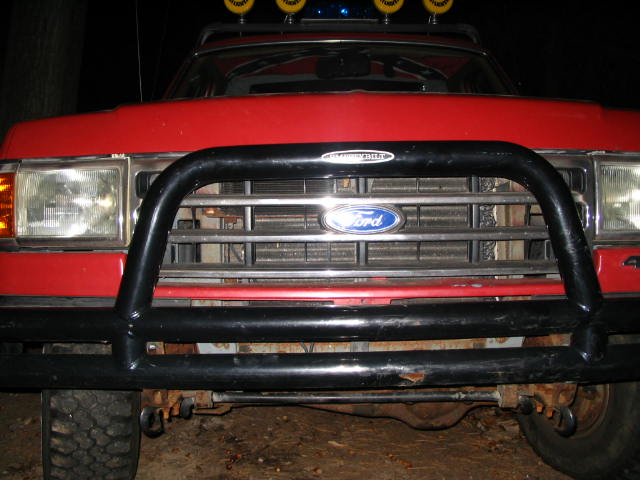 Front grille of a red Ford truck at night with black tube brush guard and light bar