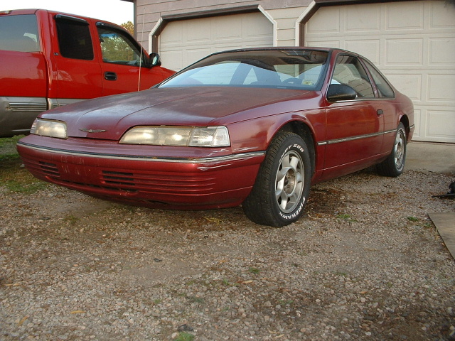 Red 1989 Ford Thunderbird coupe parked outside, with a red pickup truck to its left