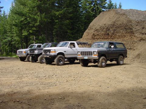 1979 Ford Bronco - Four Ford Broncos, including a 1979 model, are parked in a line on a dirt lot with trees and a pile of gravel in the background.