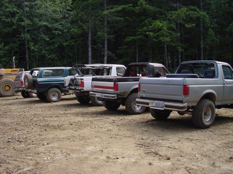 1979 Ford Bronco - Multiple off-road vehicles are parked in a line on a dirt lot, with a forest in the background.