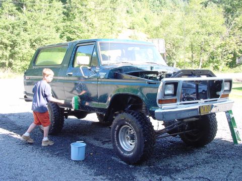 1979 Ford Bronco - A boy washes a green 1979 Ford Bronco in a driveway with trees in the background.