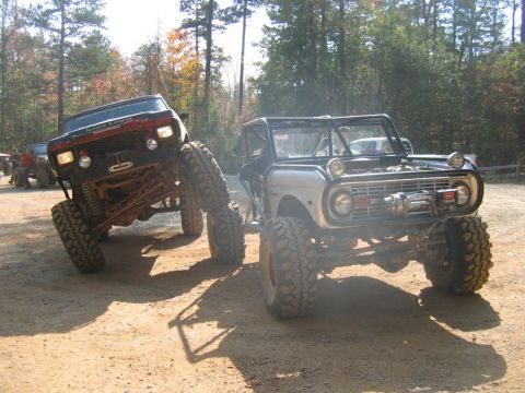 1992 Ford Mustang - Two custom off-road vehicles are parked on a dirt trail, with one vehicle lifted high on its suspension.