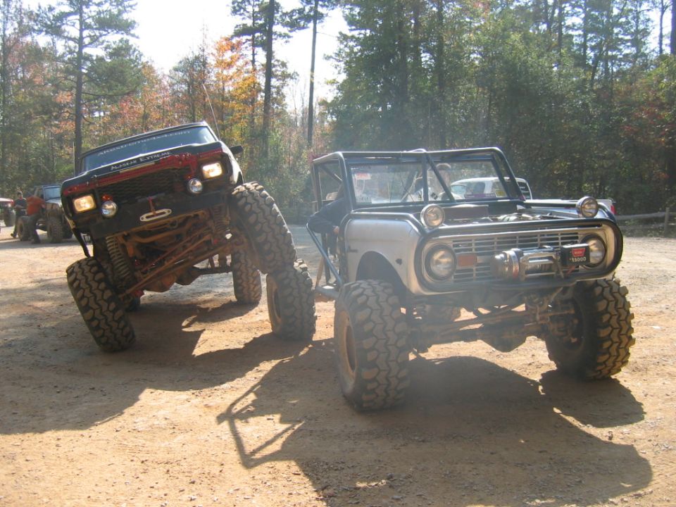Two heavily modified SUVs, one silver Bronco and one red Jeep-like vehicle, parked on a dirt path with trees in the background