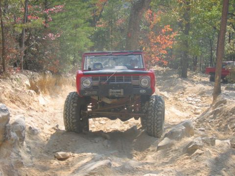 1992 Ford Mustang - A red off-road vehicle navigates a challenging rocky trail with trees and fall foliage in the background.