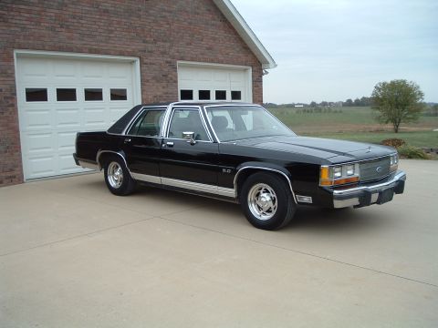 1989 Ford Crown Victoria LTD - A black sedan with chrome accents sits in a driveway in front of a residential garage.