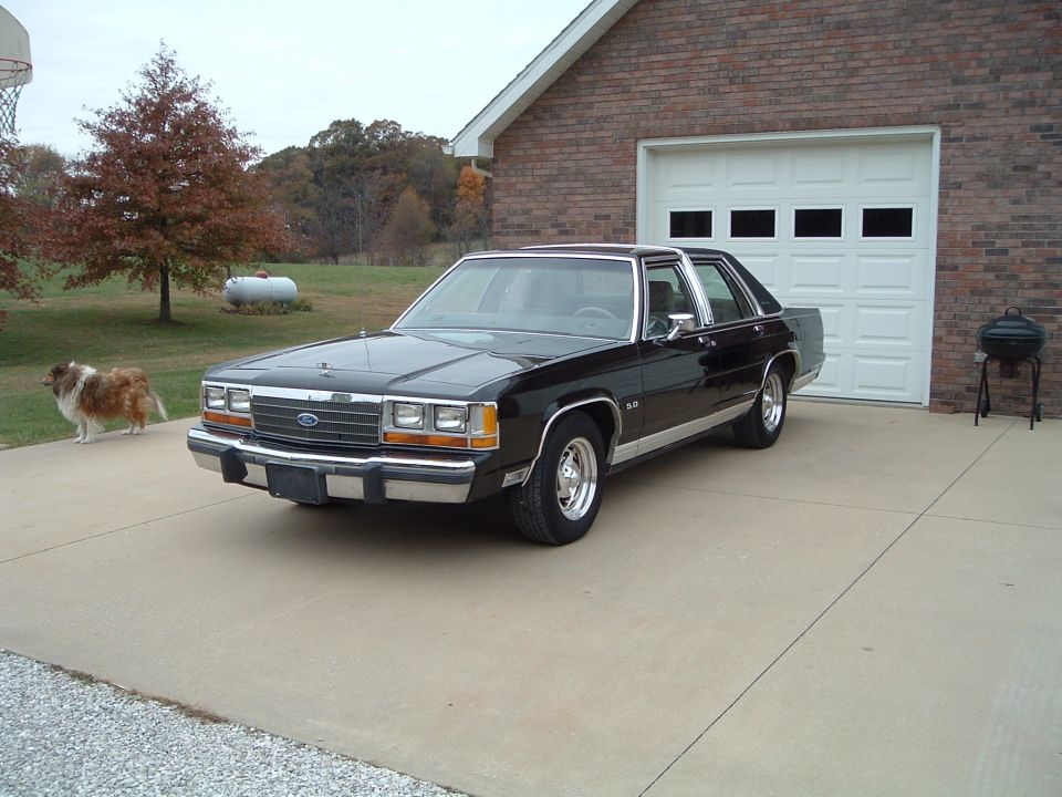 Black 1980s Ford sedan, 3/4 front view on driveway by brick garage, chrome grille and 5.0 badge
