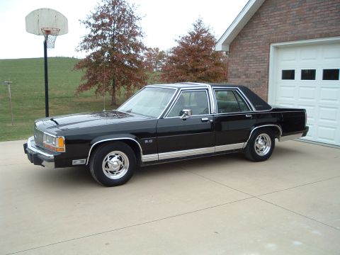 1989 Ford Crown Victoria LTD - This black sedan, likely from the 1980s, is parked in front of a brick house and garage.