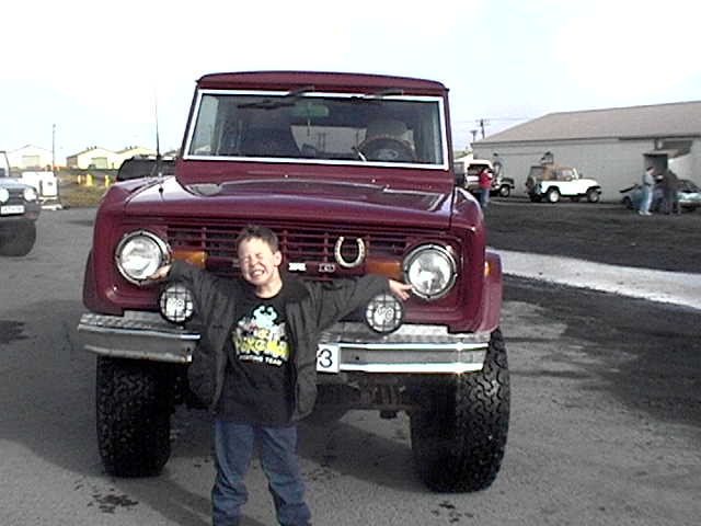 Boy posing with arms out in front of a maroon lifted early Ford Bronco with big tires and bumper lights
