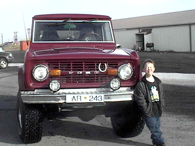Maroon early Ford Bronco, front view, with boy standing beside it on a dirt lot