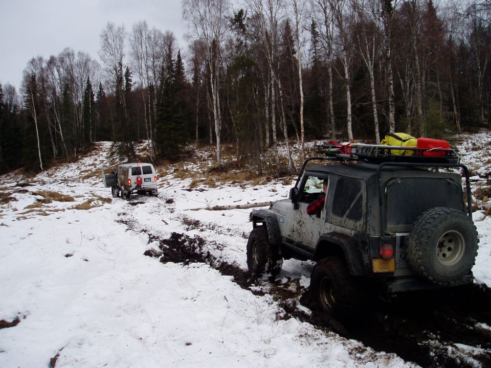 Two off-road vehicles driving through mud and snow in a wooded area. The lead vehicle is a white van, and the trailing vehicle is a silver Jeep with a roof rack