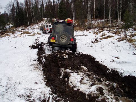 2000 Ford E350 - Two off-road vehicles navigate a challenging muddy and snowy trail in a wooded area.