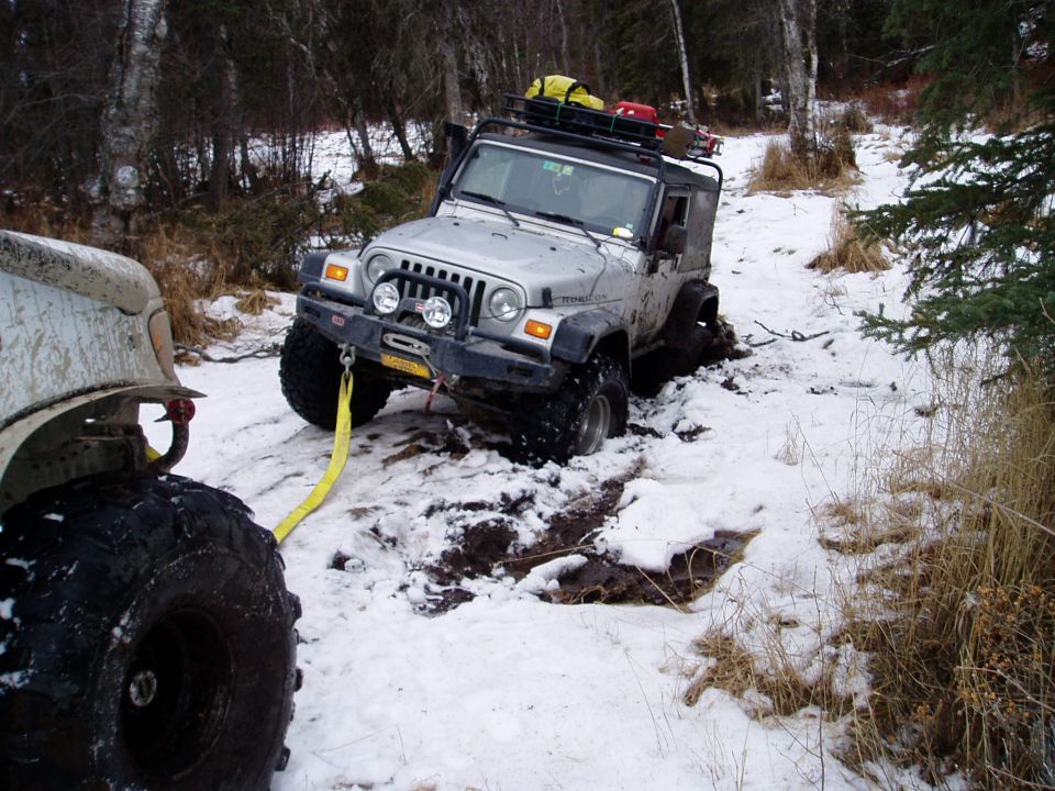 Silver Jeep Wrangler stuck in snow, yellow tow strap attached to front bumper, being pulled by an unseen vehicle