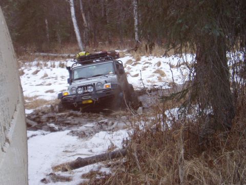 2000 Ford E350 - A Jeep Wrangler struggles through a muddy, snow-covered trail in a wooded area.