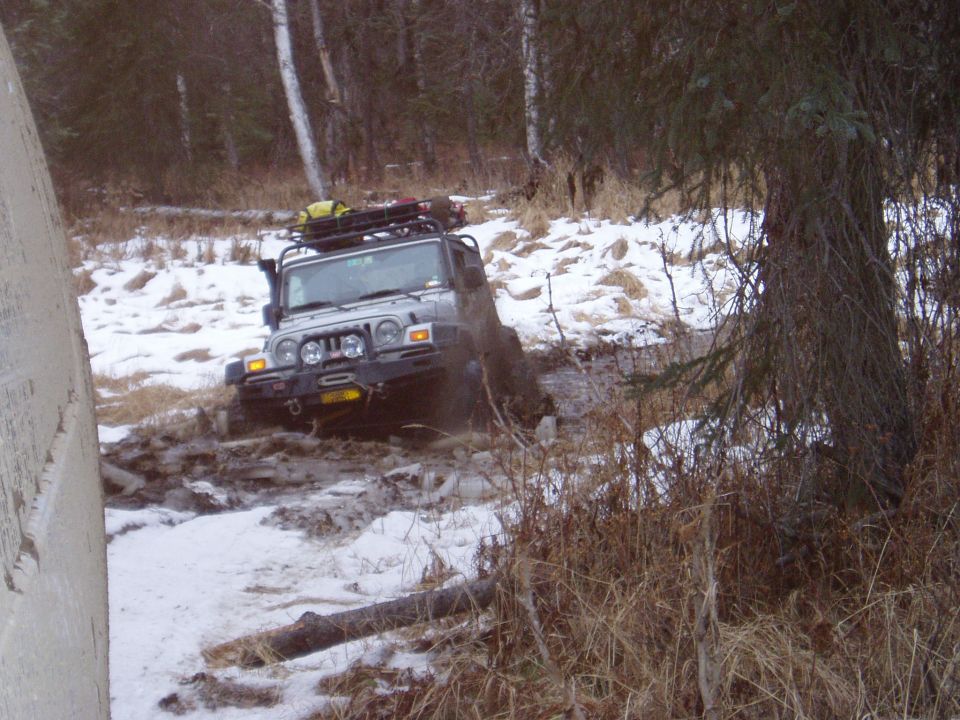Silver Jeep Wrangler navigating a muddy trail with snow on the ground