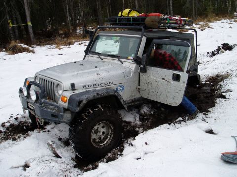 2000 Ford E350 - A Jeep Wrangler is stuck in a muddy and snowy off-road trail, with its door open and a person inside.