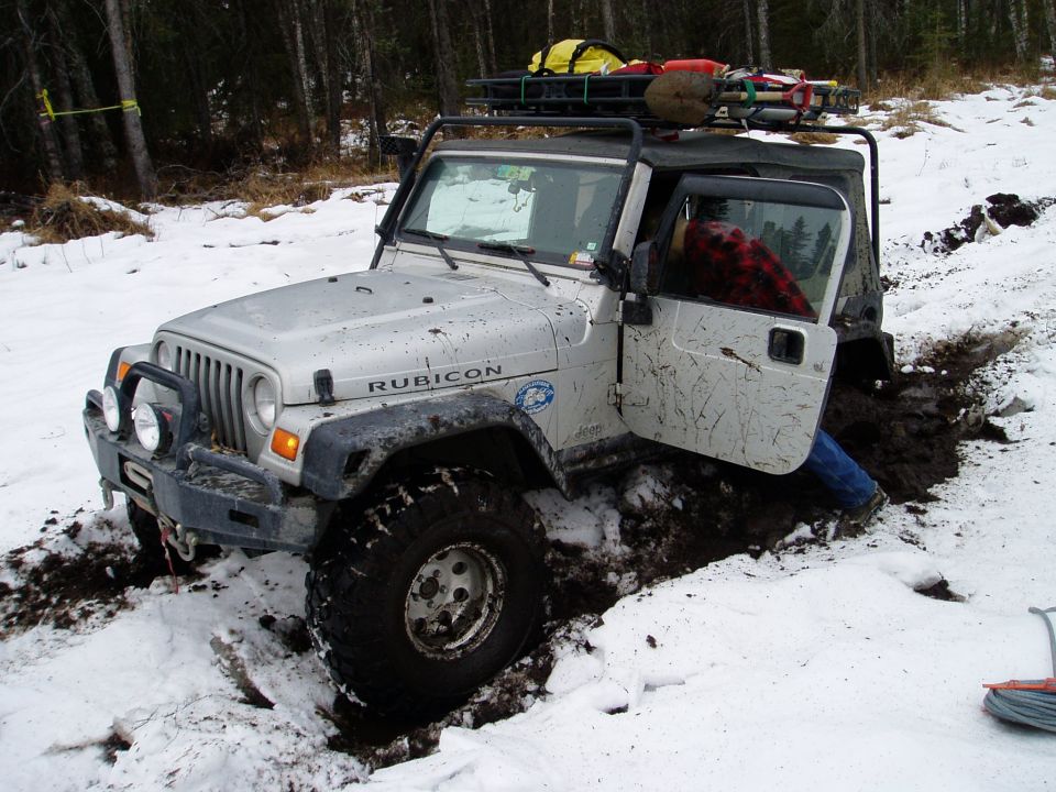 Silver Jeep Wrangler Rubicon stuck in a muddy ditch with snow on the ground. The driver's door is open and a person in a red plaid shirt is visible inside
