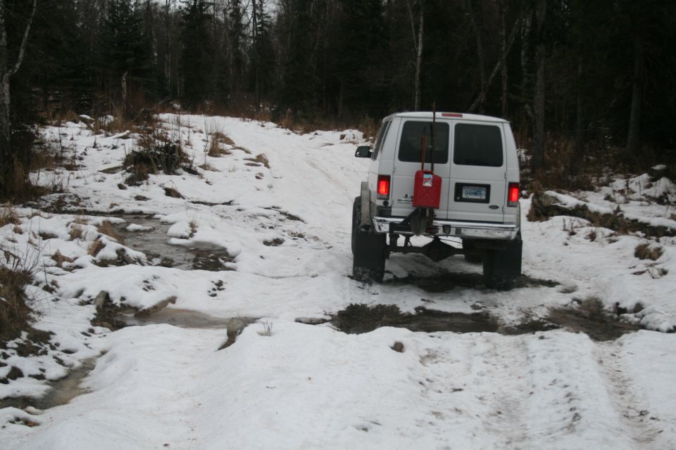 White van with a red gas can mounted on the back drives on a snow-covered trail through woods