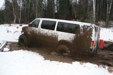 2003 Jeep TJ - A white van is stuck in a muddy, snowy off-road trail, with mud spraying from its wheels.