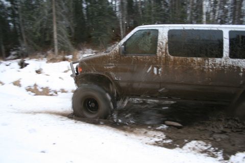 2003 Jeep TJ - A van is driving through a muddy and snowy terrain, with trees visible in the background.