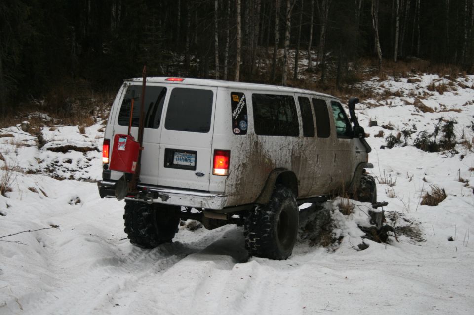 White van with big tires and a shovel and gas can on the back, driving in snow
