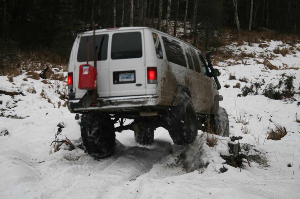 White van with large tires driving through snow, red gas can mounted on rear