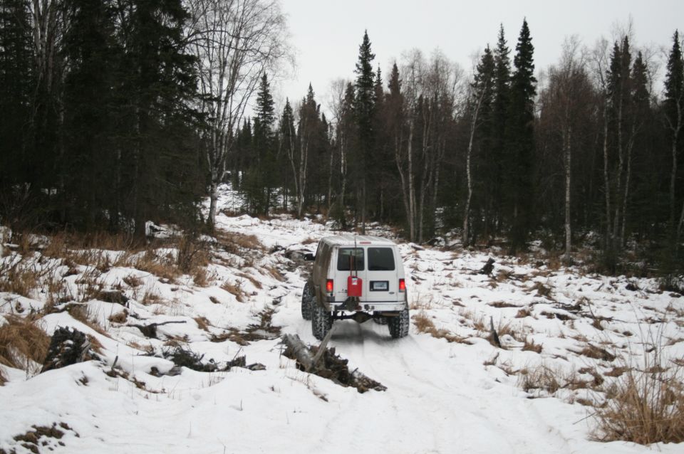 White van with a red gas can mounted on the rear bumper drives up a snow-covered forest trail