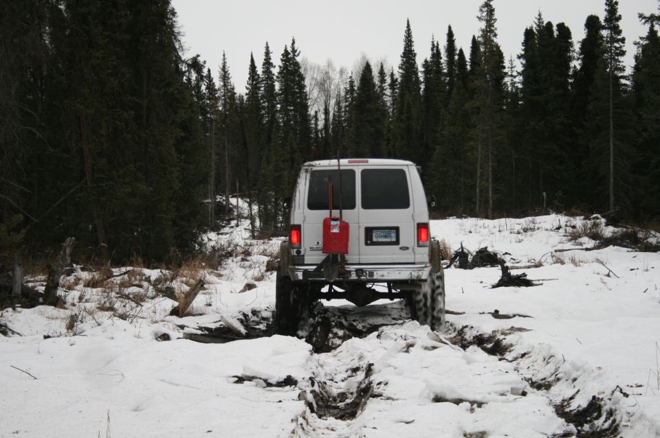 White van with a red gas can on the back is stuck in a muddy, snowy rut in a forest