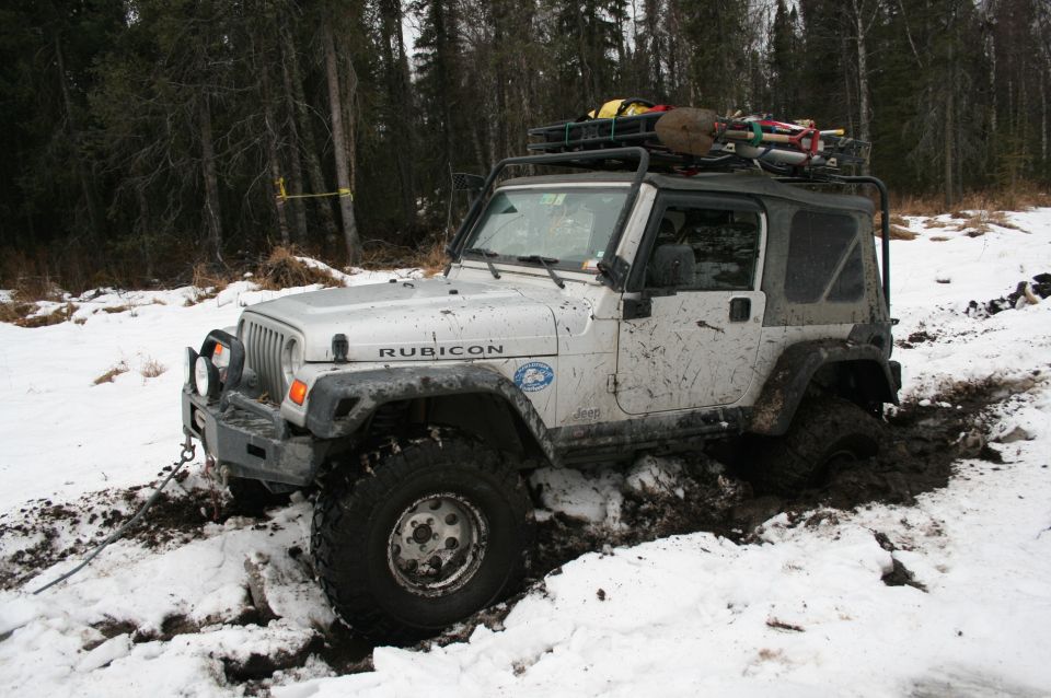 Silver Jeep Wrangler Rubicon stuck in mud and snow, with a roof rack loaded with gear