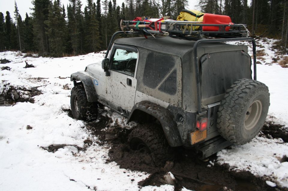 White Jeep Wrangler stuck in mud and snow, with roof rack loaded with gear