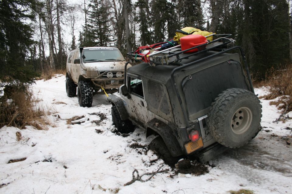 Mud-splattered 2003 Jeep TJ stuck in snow, being pulled by a lifted Ford Explorer