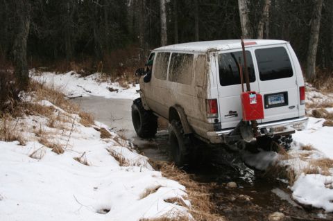 2003 Jeep TJ - A white van with off-road tires drives through a shallow stream in a snowy, wooded area, carrying a red gas can on its rear bumper.