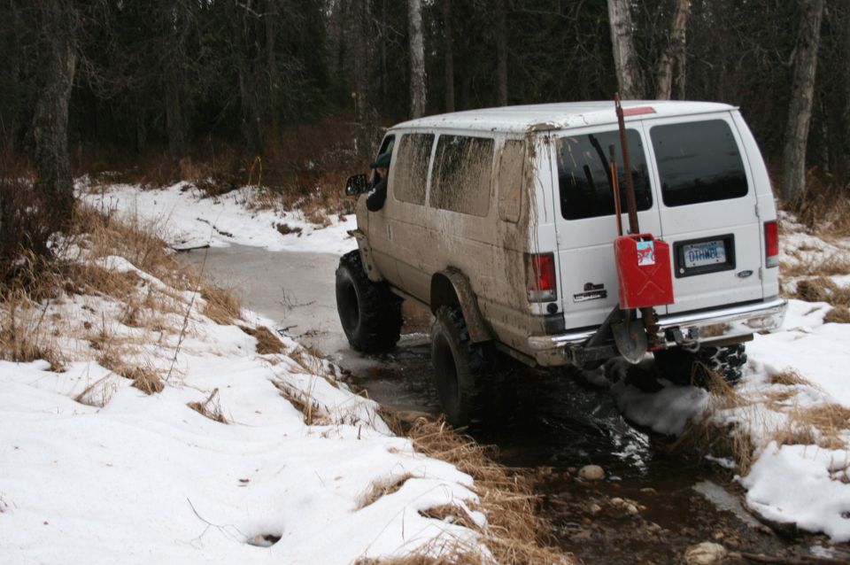 White van with large tires navigating a snowy stream, red gas can mounted on rear