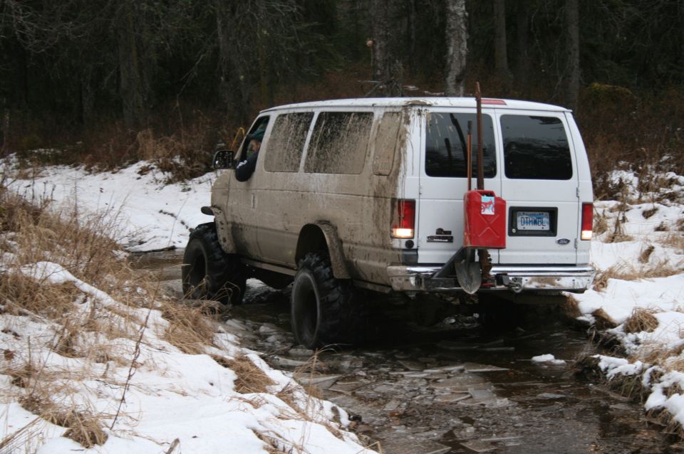 White van with big tires driving through a snowy, muddy trail. Red gas can and shovel mounted on the back