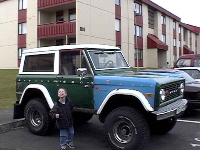 Green and blue early Ford Bronco with white top on all-terrain tires in a parking lot