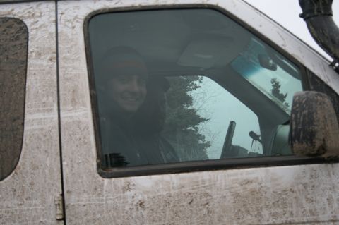 2003 Jeep TJ - Man smiles from inside a mud-covered vehicle, seen through the driver's side window.
