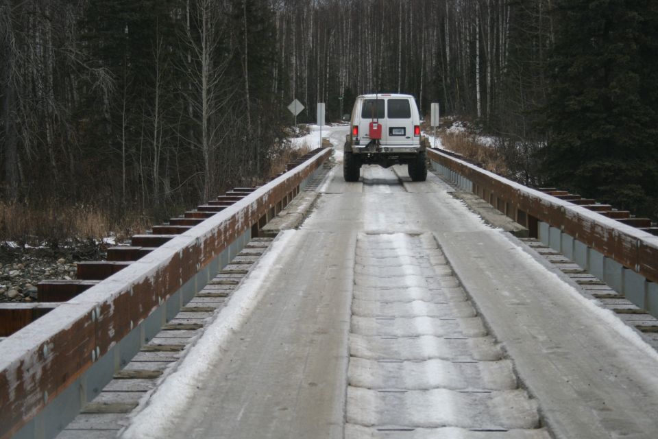 White van driving on a snow-covered bridge with trees in the background