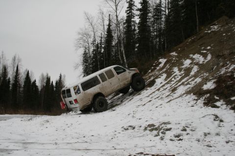 2003 Jeep TJ - A white van with oversized tires navigates a snow-covered incline next to a forest.