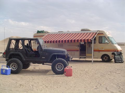 2000 Jeep TJ - Jeep TJ and RV camping setup in a sandy, desert-like environment under a cloudy sky.