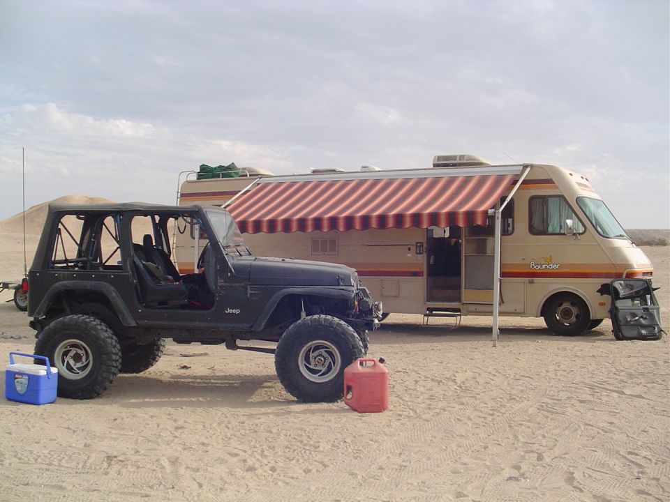Black Jeep TJ with aftermarket wheels and tires, parked next to a tan RV with red striped awning. Red gas can and blue cooler nearby