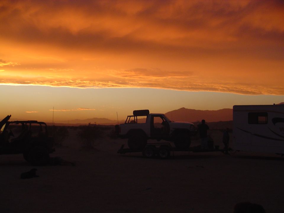 White Jeep Wrangler on a trailer at sunset, with a camper and another off-road vehicle visible