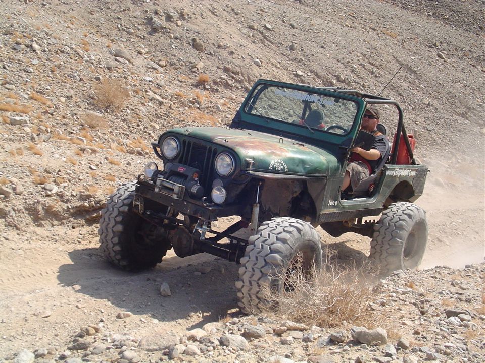 Green Jeep CJ-7 off-roading on a dirt path, front-left 3/4 view. Modified with large tires, winch, and roll cage